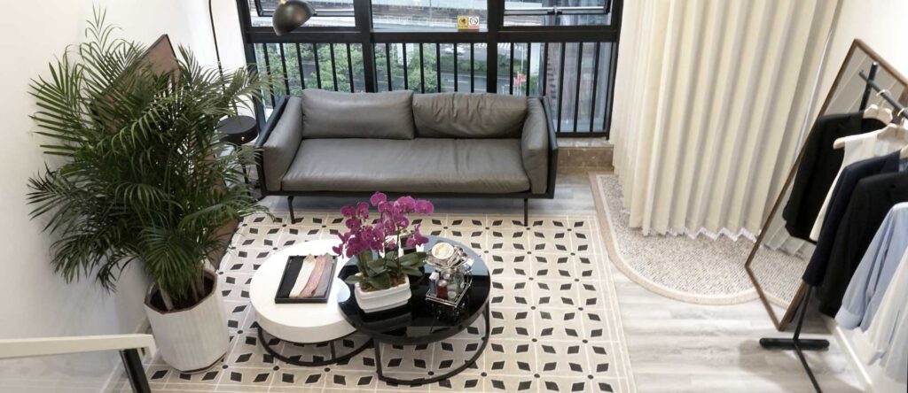 Modern living room with a gray sofa, round coffee table, potted plants, books, and a balcony view, featuring geometric patterned rug and minimalist decor.