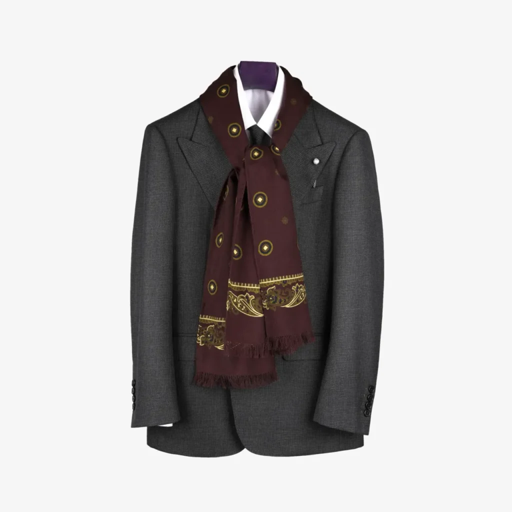 A gray suit jacket with a white dress shirt and the Sarava – Wool and silk scarf in Burgundy draped over the collar creates a stylish ensemble, showcasing the elegant scarf as an accessory, all displayed neatly on a hanger.