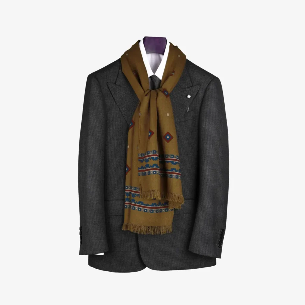 A dark gray suit jacket with peaked lapels and a white shirt is displayed on a stand, elegantly finished with the Kandora – Wool and Silk Scarf in Mustard, stylishly tied at the neck.