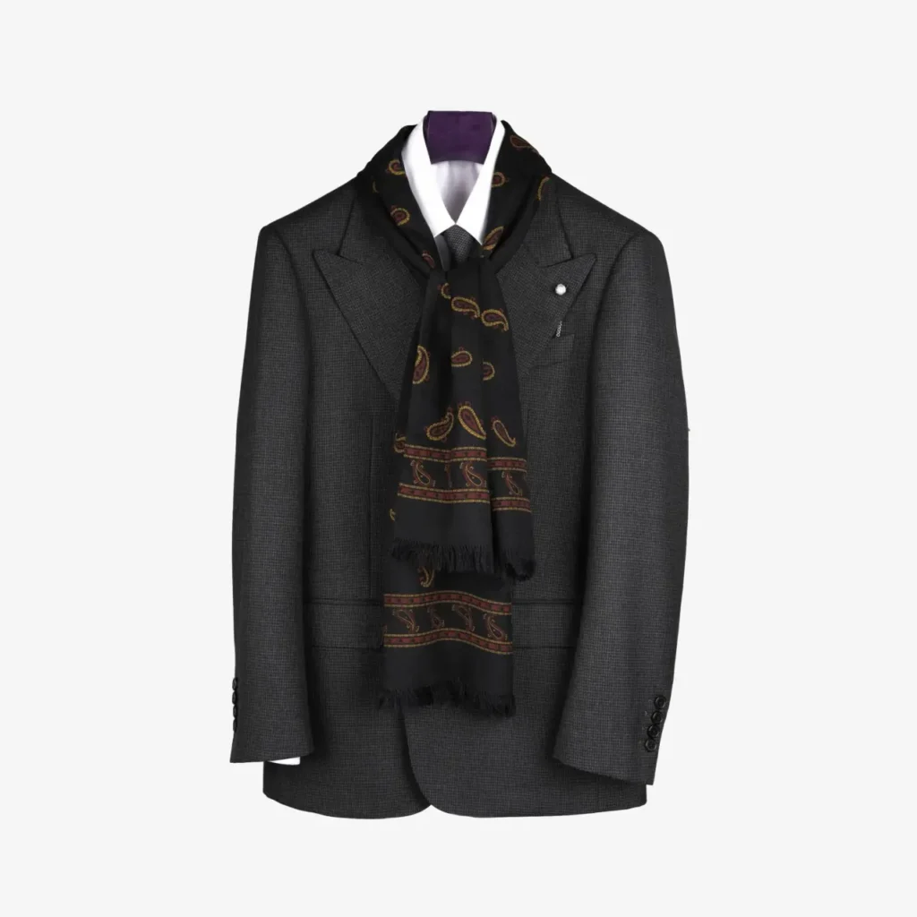 A dark gray suit jacket with a white dress shirt and the Lunema wool and silk scarf in indigo, featuring a luxurious gold paisley pattern, draped around the collar and displayed on a hanger.