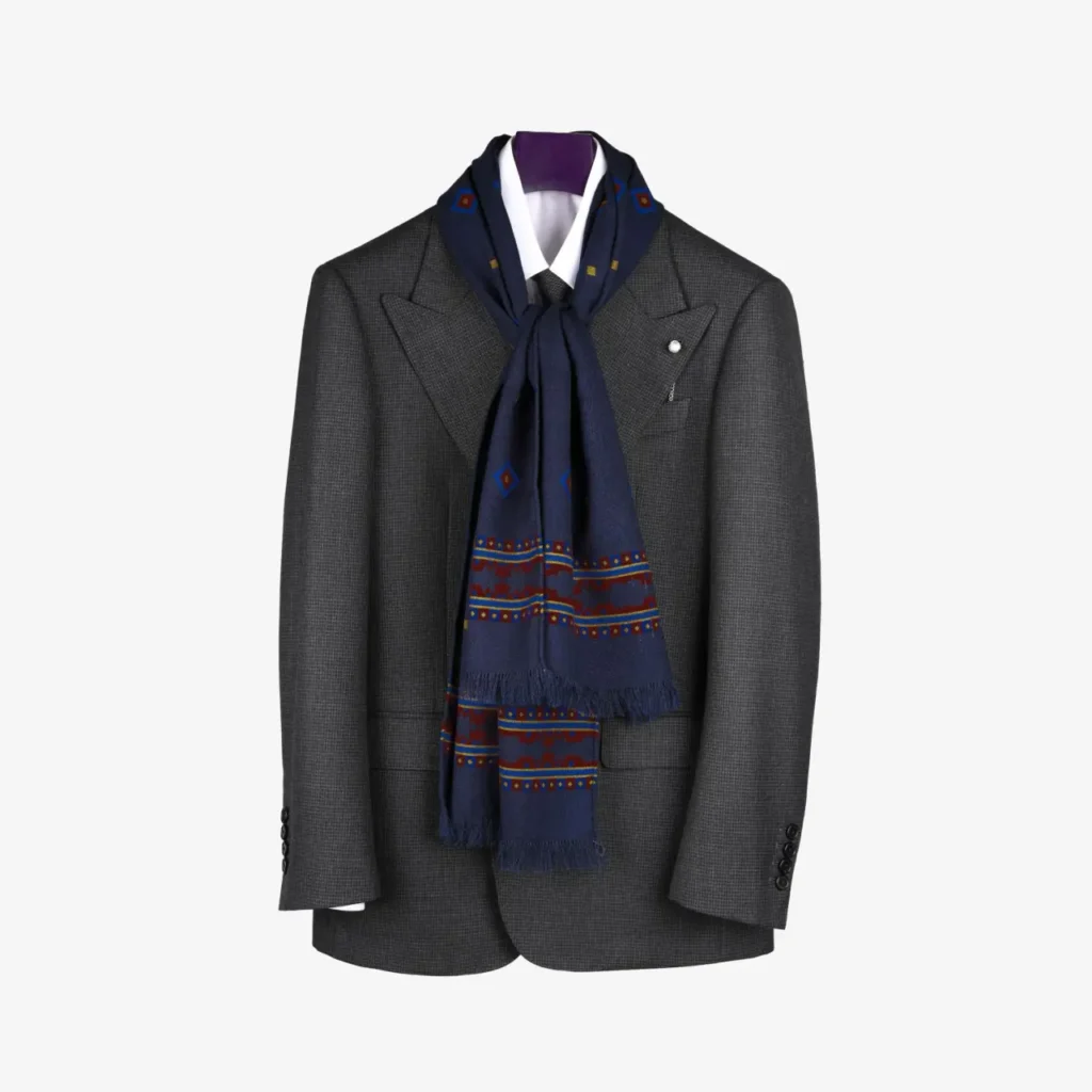 A gray suit jacket with peaked lapels is displayed on a hanger, featuring the Kandora – Wool and Silk Scarf in Navy draped around the collar over a crisp white dress shirt.