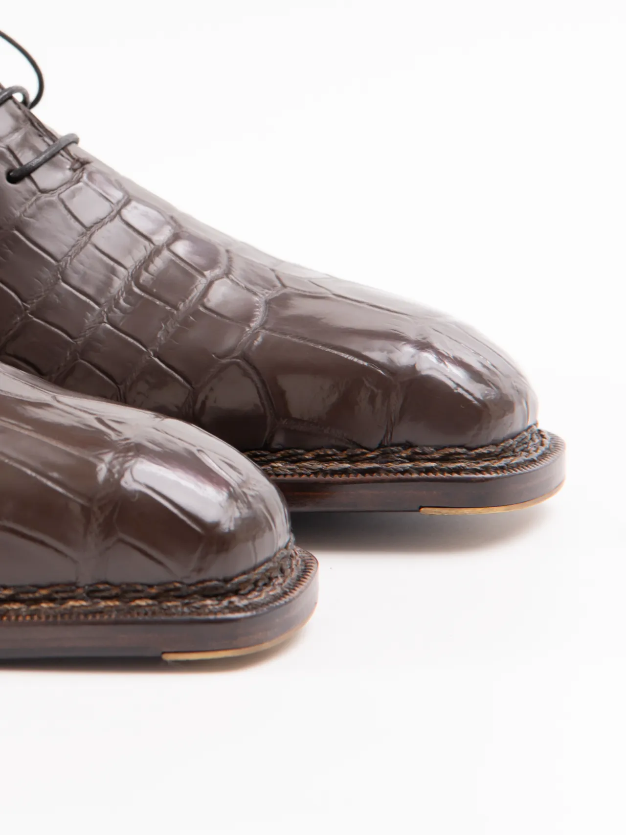 Close-up of the front portion of dark brown, crocodile-patterned leather dress shoes with thick soles on a white background.